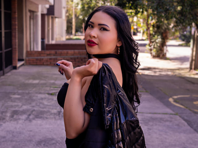 OliviGrace with dark hair, red lipstick, black top, and shiny black jacket, holding it off her shoulder, looking over her shoulder with a slight smile, showing her ass.