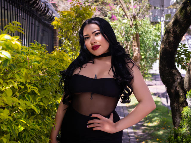 OliviGrace with black curly hair, red lipstick, and a sheer black top showing her tits, standing in a sunny garden with green foliage and a black fence.