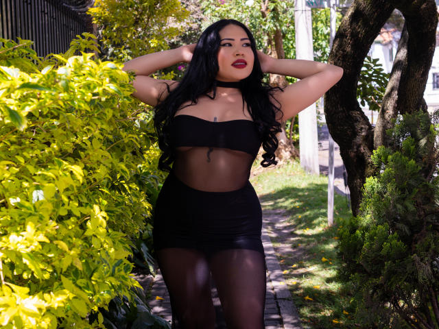 OliviGrace with long black hair, dark red lipstick, wearing a black sheer top and tight black skirt, standing with hands in hair, sexy, curvy, and showing her ass.
