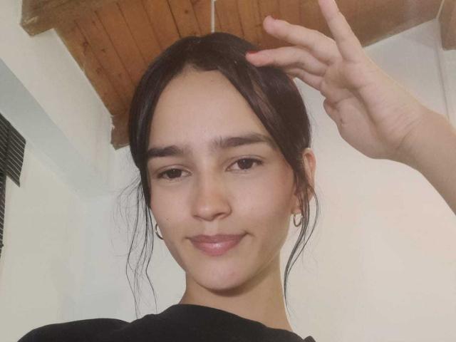 MelodyMansson has dark hair in a loose bun, light skin, small hoop earrings, and is making a peace sign with a slight smile, wearing a black shirt.