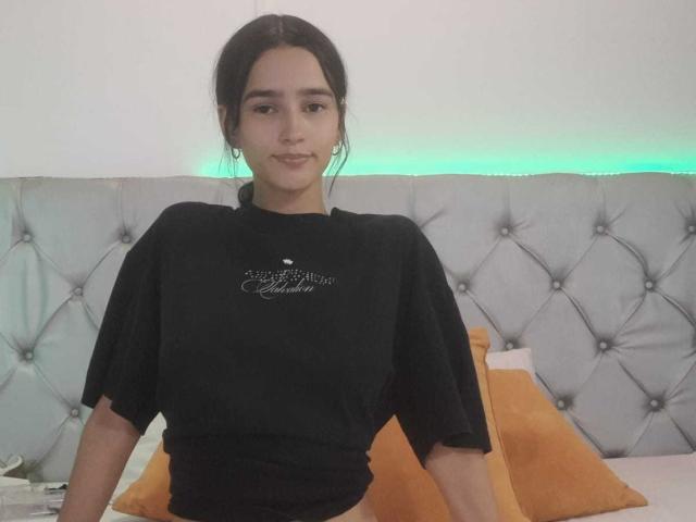 MelodyMansson, with dark hair in a ponytail, wearing a black shirt, sits on a bed with orange pillows, looking at the camera, smiling slightly, in a bedroom.