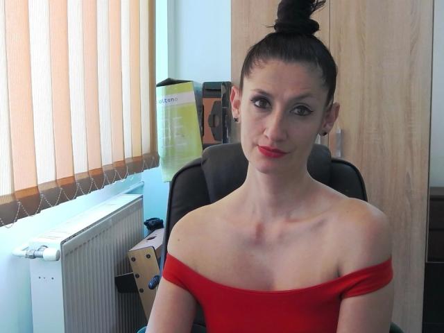 BeatrixCharmm with black hair in a bun, wearing a red off-shoulder top, showing small tits and a slim build, sitting in a chair with a slight smile.