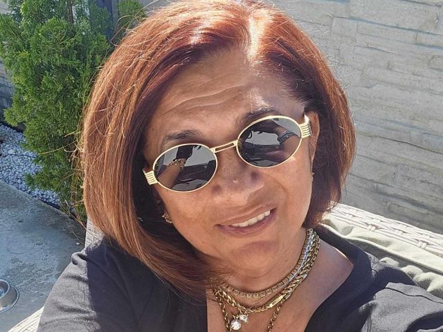 ClassybutNaughty with red hair, wearing gold sunglasses and multiple necklaces, smiling at the camera. Her tits are partially visible under a black top. Sunlit outdoor setting.