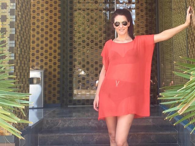 JoliDanielle, wearing a sheer red dress, shows off her tits with a V-cut, standing with one arm raised, smiling, wearing reflective sunglasses, in front of a tiled door.