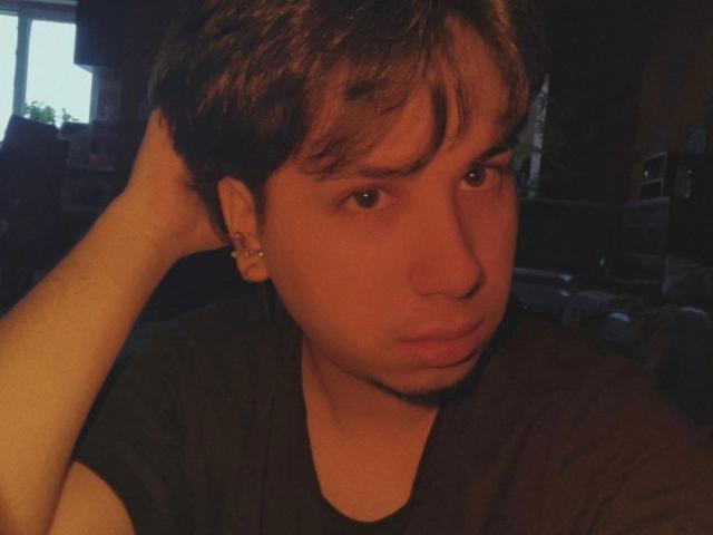 FemCupid has medium brown hair, a slight mustache, and is looking at the camera with a serious expression, hand in hair, wearing a dark shirt in dim lighting.