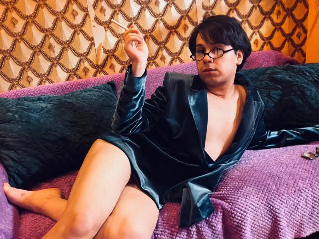 FemCupid with short black hair, glasses, and a black velvet robe, showing a deep V-neck, sitting on a purple bed, legs exposed, hand raised.