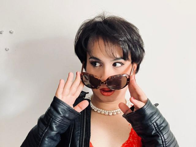 FemCupid with short black hair, wearing a black leather jacket, red lace top, pearl necklace, and large brown-tinted sunglasses, hands adjusting glasses, looking surprised.
