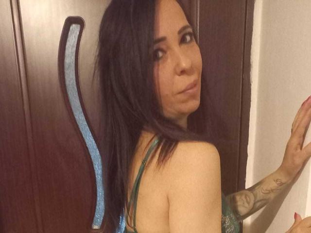 LauraSin with long dark hair, green lingerie, tattoo on right arm, looking over shoulder, smiling, brown wooden door with blue curved handle in background.