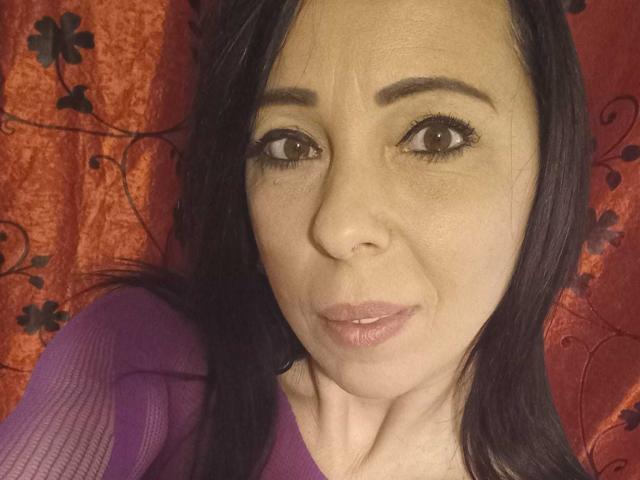 LauraSin has a pouty face, brown eyes, and black hair. She's wearing a purple top, and her tits are slightly visible. Her ass is not in the frame.