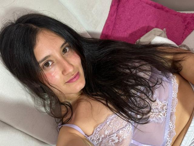 KimyRosse with long black hair, light brown eyes, and pink cheeks, wearing a lilac lace bra, lying on a white pillow, pink blanket in background.