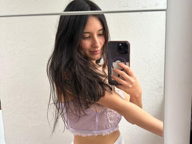 KimyRosse with long black hair, smiling, taking a mirror selfie in a lilac lace bra, holding a phone with a purple case, showing her tits and a tattoo.