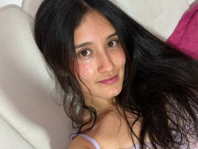 KimyRosse has long black hair, brown eyes, pink lips, and a slight smile, sitting on a white couch with a pink pillow, wearing a purple tank top.