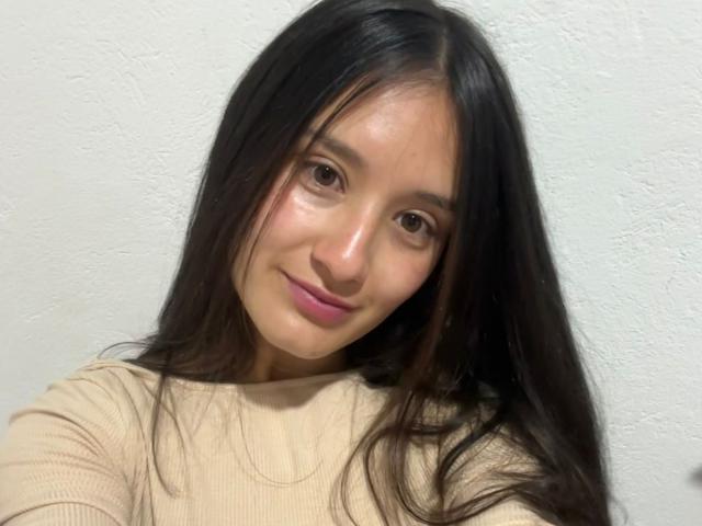 KimyRosse has long black hair, brown eyes, and pink lips. She's wearing a beige ribbed shirt, smiling slightly, and has a fair skin tone.