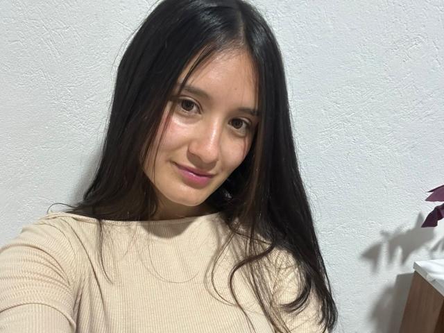 KimyRosse with long black hair, wearing a beige ribbed shirt, smiling slightly, showing subtle pink lipstick, and cute brown eyes. Her small tits are partially visible.
