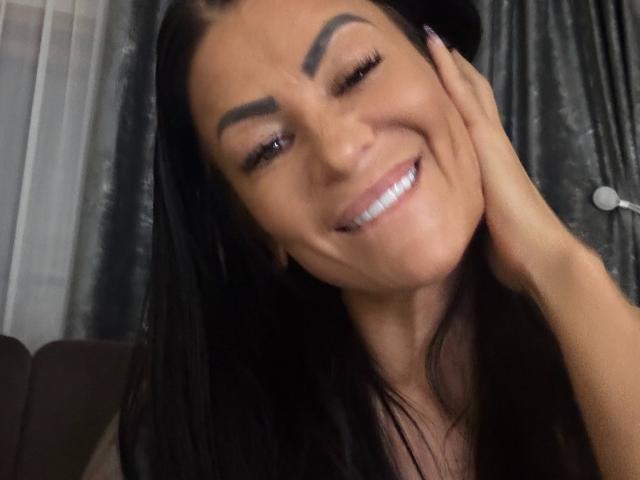 AndraD with black hair, smiling, winking, and touching her cheek. She has thick eyebrows, long eyelashes, and is wearing a black top. Her teeth are visible.