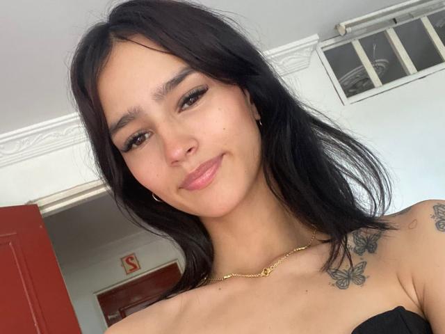 MelodyMansson has long black hair, dark eyebrows, and a slight smile. She's wearing a gold necklace and has butterfly tattoos on her shoulders. Her lips are glossy.