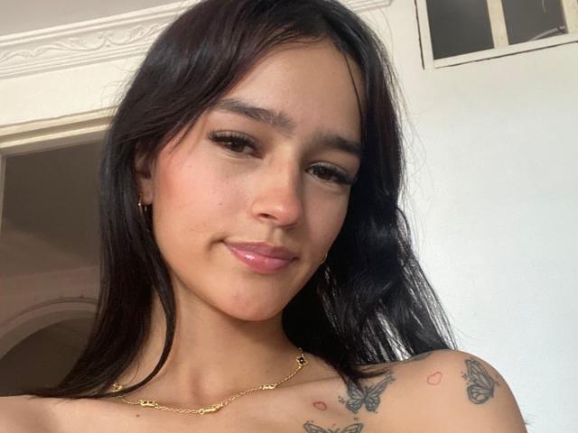 MelodyMansson has long dark hair, brown eyes, and a slight smile. She's wearing a gold necklace and has tattoos on her shoulder with butterflies and hearts.