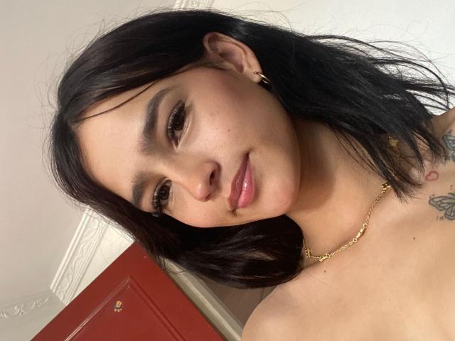 MelodyMansson has dark black hair, a slight smile, and glossy pink lips. She's wearing a gold necklace, has a tattoo, and a gold hoop earring. Her tits are visible.
