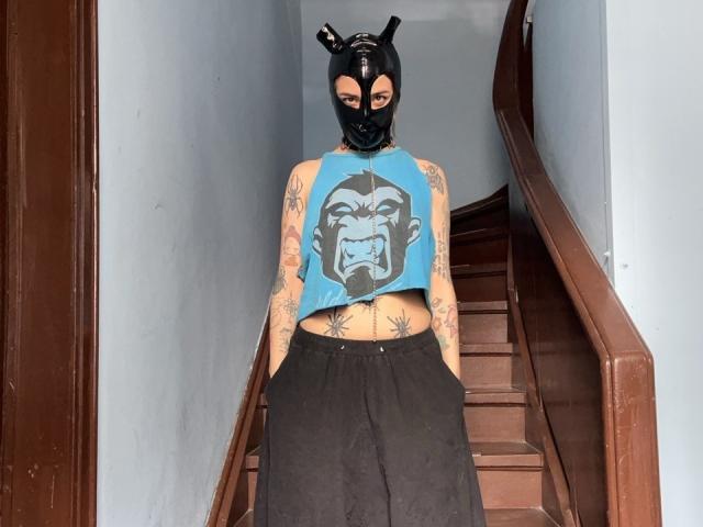 AmySpider with tattoos, blue crop top, black mask with horns, black skirt, hands in pockets, standing on stairs, showing belly tattoo, tattooed arms, and blue top with comic face.