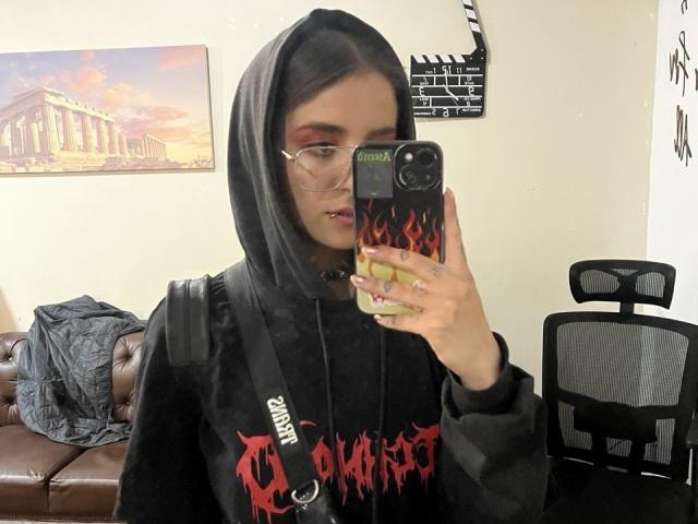 AmySpider with glasses, black hood, and "Slayer" hoodie, holding phone with flame case, taking selfie, brown leather couch, black chair, and movie camera in background.