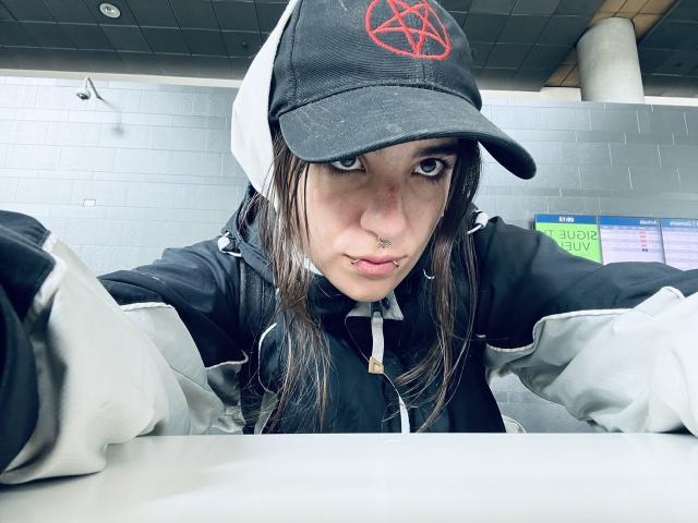 AmySpider with a black and white cap, pentagram on the front, black and grey jacket, nose ring, and lip piercing, looking at the camera with a serious expression.