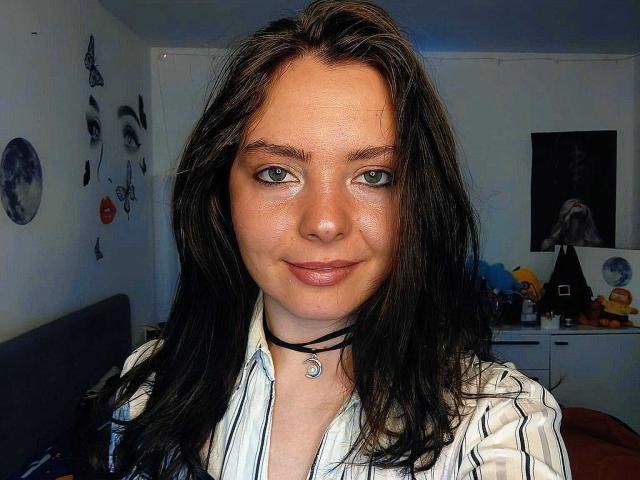 HottWetPussy69 tem cabelos pretos longos, olhos verdes e um sorriso discreto. Ela está vestindo uma camisa listrada com um colar preto e um pequeno pingente. Sua pele é clara..