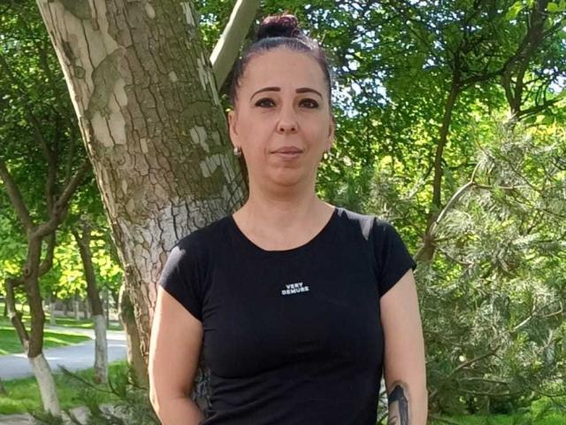 LauraSin with medium tits in a black shirt, standing in a park, has dark hair in a bun, wearing earrings, and looking slightly pouty. Tree behind her.