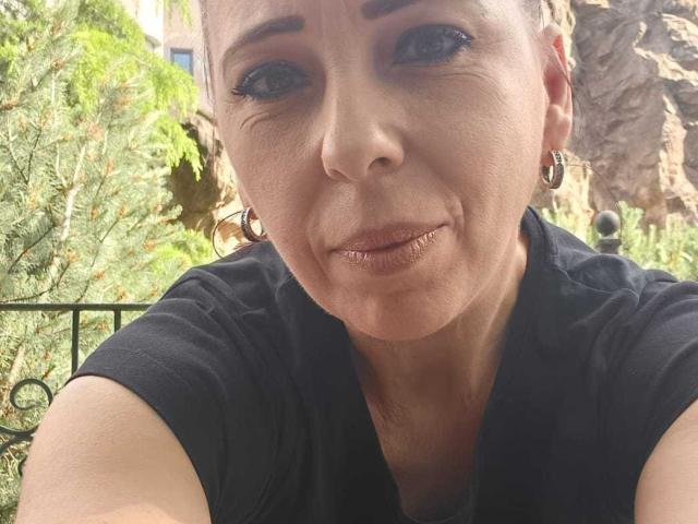 LauraSin's face with pierced nose, dark eyeliner, and black shirt, showing her upper tits and slightly parted lips with a subtle smile. Her skin is light, and she's outside.