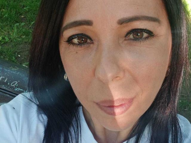 LauraSin's face with dark brown eyes, black eyeliner, and subtle pink lipstick, wearing a white shirt, with straight black hair and small earrings, in bright sunlight.