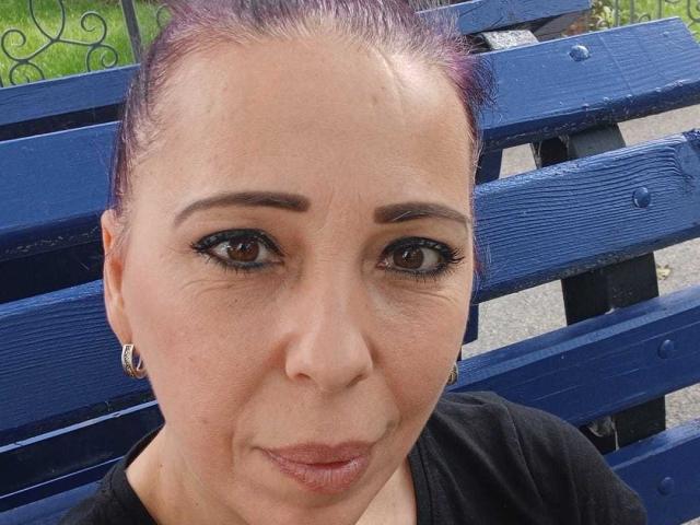 LauraSin's face, with purple-tinted hair, black eyeliner, and small hoop earrings, sitting on a blue wooden bench. She has a slight smile and a black shirt.