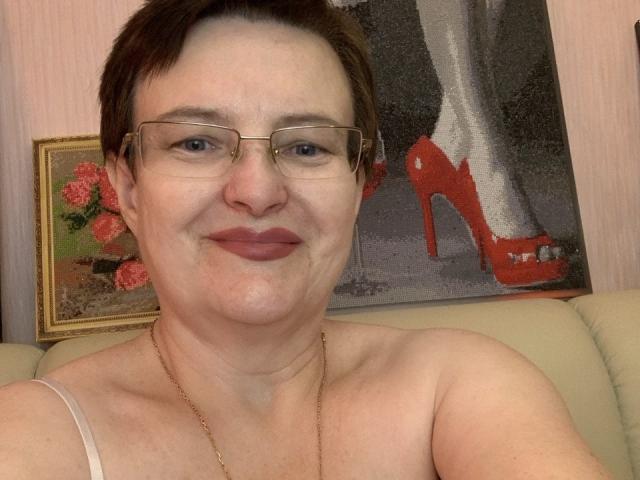 MargieEvans with short brown hair, glasses, and light pink lipstick, smiling, showing her bare shoulders, wearing a thin gold necklace, and a beige strap top, with red high heels.