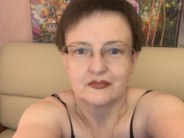 MargieEvans with short brown hair, glasses, and red lipstick, wearing a black spaghetti strap top, showing her face and upper chest. Her tits are small, and she has a gold necklace.