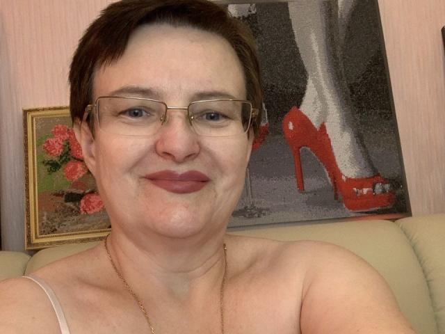 MargieEvans with short brown hair, glasses, and a subtle smile, wearing a gold necklace and a strapless top, showing her tits, in a picture with red high heels.