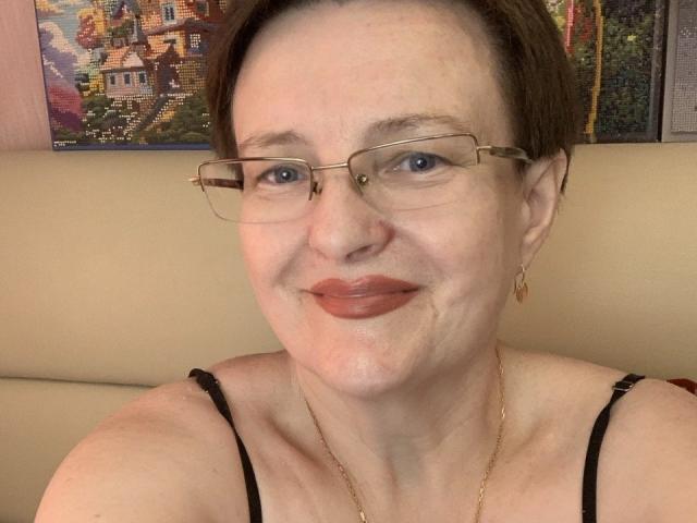 MargieEvans has short brown hair, glasses, and is smiling with her lips slightly parted. She's wearing a black strap top and a gold necklace. Her tits are small.