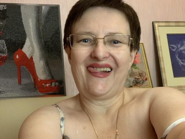 MargieEvans with short brown hair, glasses, and a smile, tongue out, showing her tits in a strapless top, wearing a gold necklace, in a room with art.