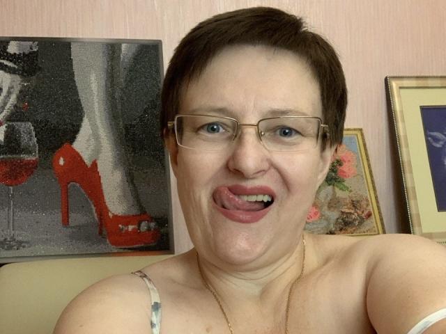 MargieEvans has short brown hair, glasses, and is sticking out her tongue. She's wearing a spaghetti strap top, showing her tits, and has a gold necklace.