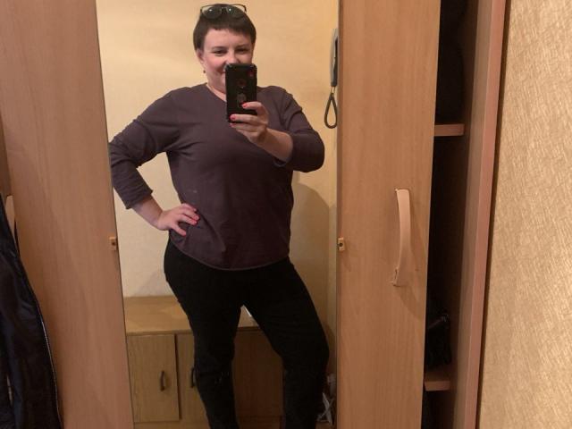 MargieEvans with short brown hair, wearing a purple long-sleeve shirt and black pants, standing in a mirror selfie, hand on hip, holding a phone. Boobs are small.