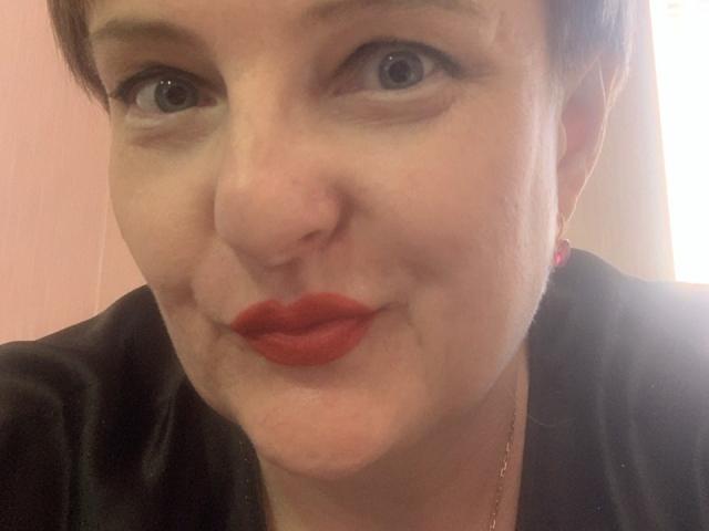 MargieEvans with short brown hair, blue eyes, and bright red lipstick, wearing a black top, has a slightly puffy face and is smiling softly at the camera.