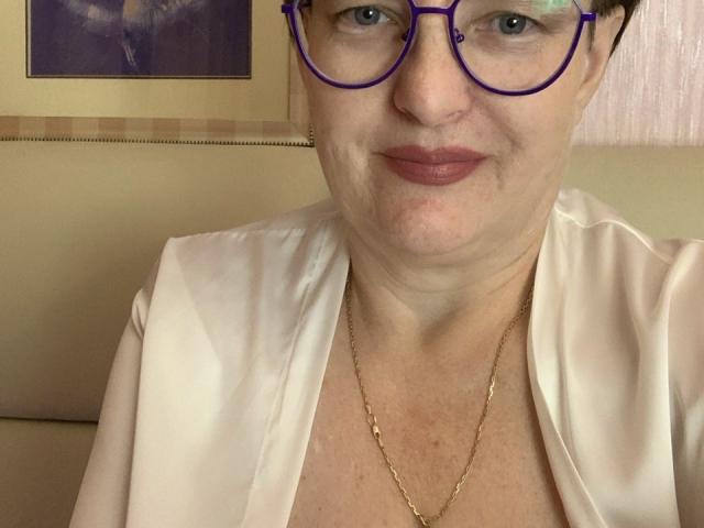 MargieEvans with purple glasses, white blouse, gold necklace, and pink lipstick, showing her face and upper chest. She has short black hair and a slight smile.