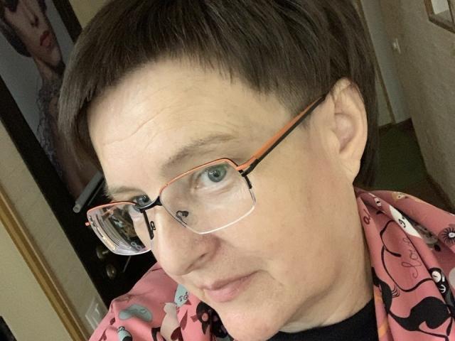 MargieEvans has short brown hair, blue eyes, and wears pink glasses with a pink shirt featuring cartoonish designs. She's smiling slightly, showing light wrinkles on her forehead.