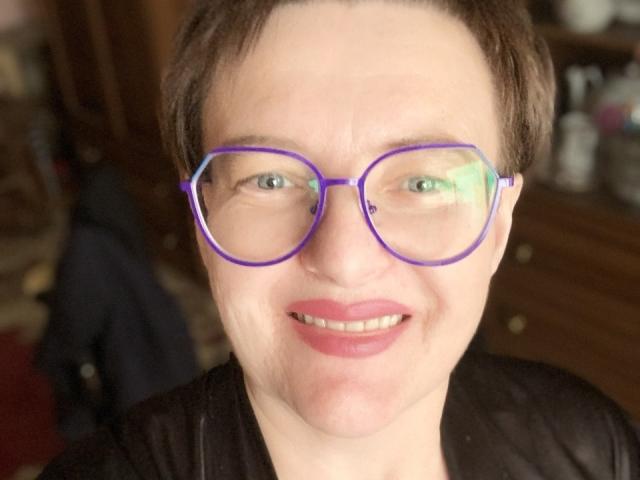MargieEvans with short brown hair, purple-framed glasses, and pink lipstick, smiling at the camera. She's wearing a black shirt, and her blue eyes are slightly squinted.