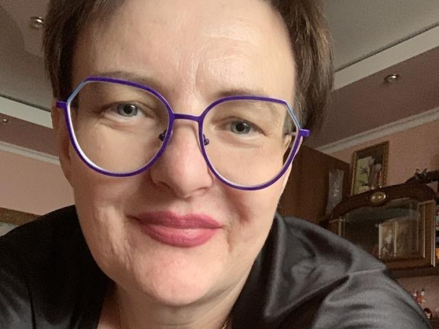 MargieEvans with short brown hair, purple-framed glasses, and pink lipstick, smiling at the camera, wearing a black shirt, showing her nose and mouth slightly sagging.