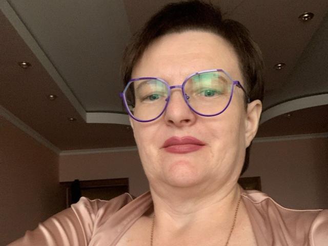 MargieEvans has short brown hair, purple glasses, and pink lipstick. She's wearing a pink satin top with a gold necklace. Her face is slightly puffy, and she's smiling.