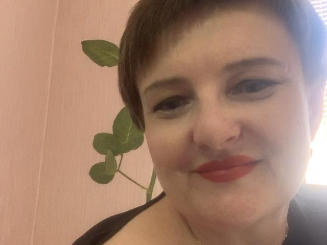 MargieEvans with short brown hair, red lipstick, and a nose ring, smiling at the camera. Green leafy plant in the background. Soft pink wall. Gentle light.