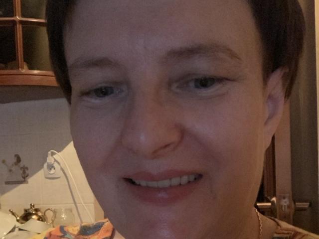 MargieEvans's face, short brown hair, blue eyes, slight smile, fair skin, small nose, subtle makeup, visible teeth, necklace, dimly lit room, soft light, casual, relaxed, slightly sweaty.