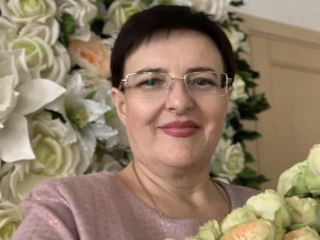 MargieEvans, short black hair, glasses, pink lipstick, pink sweater, holding bouquet of white and pink roses, slightly smiling, beige wall in background.