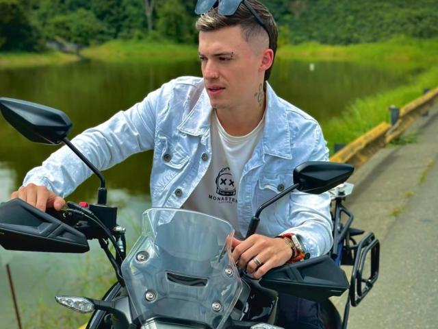 ErosHot with short blond hair, wearing a white denim jacket and white shirt, riding a black motorcycle with a clear windshield, hands on the handlebars.