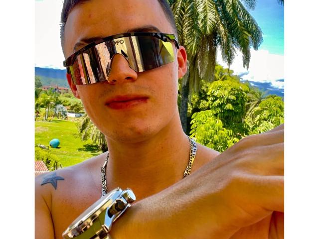 ErosHot with reflective sunglasses, nose ring, and chain necklace, shirtless, showing a tattoo on the right shoulder, holding a metal bracelet, palm tree in the background.