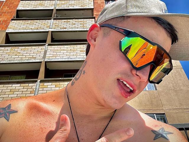 ErosHot with a shirtless, tattooed, light-skinned, muscular build, wearing a gray cap and reflective rainbow sunglasses, showing a finger and a partially open mouth.