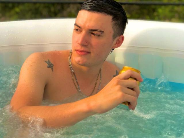 ErosHot, shirtless, with short dark hair, holding a yellow can in a bubbling hot tub, has tattoos on his right shoulder and a chain necklace, looking slightly bored.
